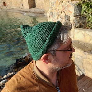 May include: A green crocheted hat with a pointed top. The hat is made of a textured yarn and has a folded brim. The hat is worn by a person wearing a brown jacket.