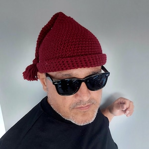 May include: A deep burgundy crochet beanie featuring a pointed top and a matching yarn tassel hanging from the peak. The hat has a thick, folded brim and a textured, hand-stitched appearance, crafted from thick yarn in a solid, dark red color.