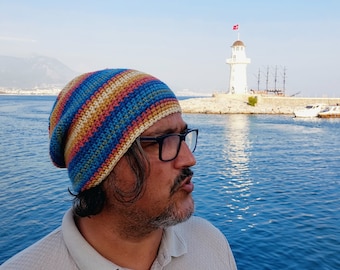May include: A man wearing a colorful crocheted beanie and glasses is standing by the water. He is looking towards a white lighthouse in the distance.