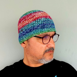 May include: A multi-colored crocheted beanie with a blue, green, pink, and red color scheme. The beanie is worn by a person with a beard and glasses.