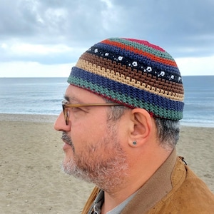 May include: A colorful, striped crochet beanie in shades of blue, brown, green, red, and tan. The hat features a geometric pattern and is worn by a person with glasses on a beach.