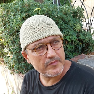 May include: A light beige crocheted beanie hat. The hat is worn by a person wearing round, tortoise-shell glasses and a black t-shirt. The background features green foliage and a wrought iron fence.