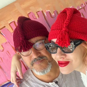 May include: Two people wearing matching red crocheted hats with tassels. One hat is a darker shade of red. The person on the right is wearing black sunglasses and red lipstick. The background features a colorful abstract painting.