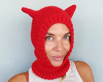 May include: A red knitted balaclava with horns. The balaclava is pulled up over the person's head, covering their ears and neck. The horns are pointed and stick up from the top of the balaclava.