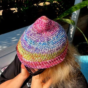 May include: A colorful crocheted beanie hat with a pointed top. The hat is made with a variety of colors, including pink, purple, blue, green, and white.