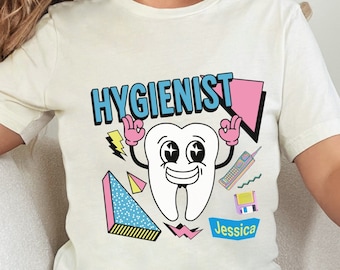 May include: Cream-colored t-shirt featuring the word "HYGIENIST" in blue and pink, with a cartoon tooth character. The tooth has a smiling face and is making a peace sign with its hands. The name "Jessica" is below the tooth.