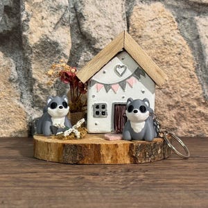 Könnte beinhalten: Eine Miniatur-Szene mit zwei grauen und weißen Husky-Figuren, einem kleinen weißen Haus mit Herz und Girlande sowie Trockenblumen, alles auf einer Holzscheibe. Ein Husky ist an einem Schlüsselanhänger befestigt.