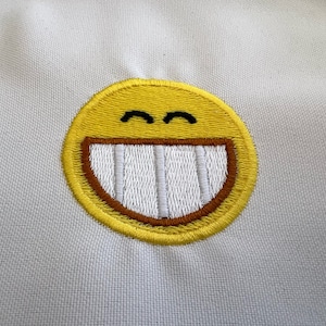 Könnte beinhalten: Ein gelbes gesticktes Smiley-Gesicht mit einem breiten, zahnigen Grinsen. Das Gesicht ist auf einen weißen Stoffhintergrund gestickt.
