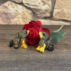 Op de afbeelding: Twee gelede vogelbeeldjes met bruine lichamen en gele voeten zijn geplaatst voor een grote rode roos. De vogels hebben zwarte accenten en staan op een houten oppervlak. Een groen blad is zichtbaar.