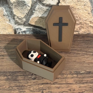 Op de afbeelding: Een miniatuurkist met een zwart kruis op het deksel en een klein figuur van een vampier erin. De vampier draagt een zwart pak en een rode vlinderdas. De kist is bruin en grijs.