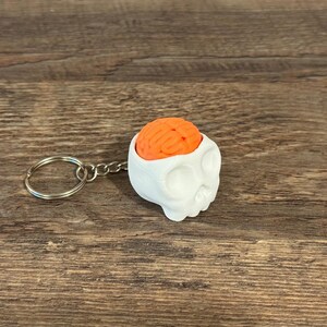 Könnte beinhalten: Ein weißer schädelformiger Schlüsselanhänger mit einem orangefarbenen Gehirn im Inneren. Der Schlüsselanhänger ist aus Kunststoff und hat einen silberfarbenen Metallring und eine Kette. Das Gehirn ist leuchtend orange.