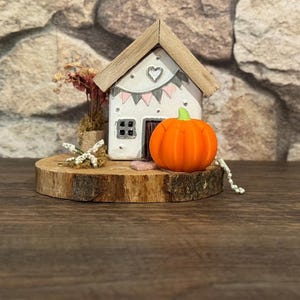Könnte beinhalten: Ein weißes Miniaturhaus mit Holzdach, Herz und Banner. Ein oranger Kürbis mit grünem Stiel steht neben dem Haus auf einer Holzscheibe. Trockenblumen und eine weiße Perlenkette vervollständigen die herbstliche Dekoration.