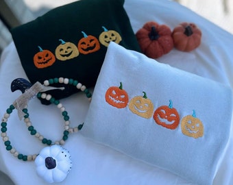 Puede incluir: Dos sudaderas con calabazas bordadas. Una sudadera es verde y la otra es azul claro. Ambas sudaderas tienen una fila de cinco calabazas con caras naranjas.