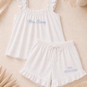 Op de afbeelding: Witte pyjamaset met een top met ruches en een short. De top is geborduurd met "Mrs. Rosey" in blauwe letters. De short heeft een taille met trekkoord en is geborduurd met "EST. 05/25/2026". De set is gemaakt van een zachte stof.