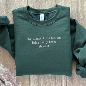 May include: A dark green sweatshirt with the text "my tummy hurts but I'm being really brave about it" in white.