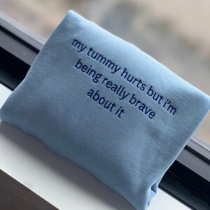 May include: Light blue sweatshirt with the text "my tummy hurts but i'm being really brave about it" embroidered in blue.