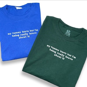 Puede incluir: Dos camisetas con el mismo texto, "my tummy hurts but I'm being really brave about it." Una camiseta es azul y la otra es verde.