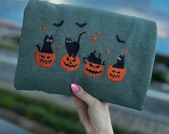 以下が含まれることがあります： ハロウィーンのデザインが刺繍されたグリーンのスウェットシャツ。デザインは、オレンジ色のジャック・オー・ランタンに座っている4匹の黒猫と、コウモリと星が特徴です。スウェットシャツは、ぼやけた青い空を背景に持っています。
