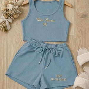 May include: A light blue ribbed knit pajama set with a tank top and shorts. The tank top has "Mrs. Raven" embroidered in gold, and the shorts have "EST 01/28/2025" embroidered in gold. A pair of white fuzzy slippers are next to the set.
