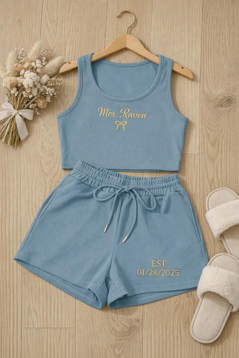 May include: A light blue ribbed knit pajama set with a tank top and shorts. The tank top has "Mrs. Raven" embroidered in gold, and the shorts have "EST 01/28/2025" embroidered in gold. A pair of white fuzzy slippers are next to the set.