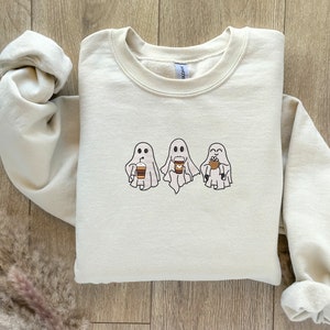 Puede incluir: Una sudadera de color crema con un gr&aacute;fico de tres fantasmas sosteniendo tazas de caf&eacute;. Los fantasmas son blancos con ojos y bocas negros. Las tazas de caf&eacute; son marrones con tapas blancas.