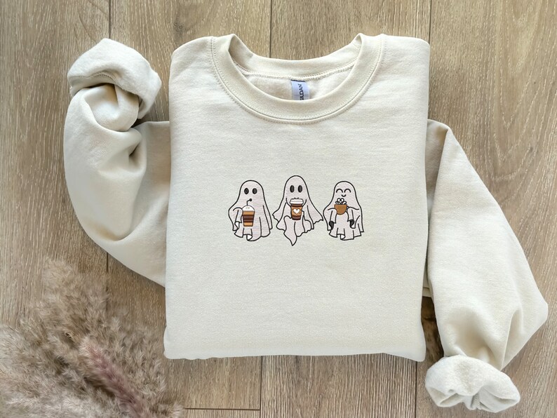 Puede incluir: Una sudadera de color crema con un gr&aacute;fico de tres fantasmas sosteniendo tazas de caf&eacute;. Los fantasmas son blancos con ojos y bocas negros. Las tazas de caf&eacute; son marrones con tapas blancas.