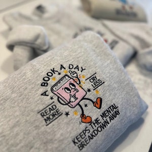 K&ouml;nnte beinhalten: Ein graues Sweatshirt mit einem gestickten Design eines Buches mit Beinen und Armen. Das Buch h&auml;lt sich einen Finger auf die Lippen und hat eine Gedankenblase &uuml;ber seinem Kopf. Der Text auf dem Sweatshirt lautet "A Book A Day Read More Cry Less Keeps The Mental Breakdown Away".
