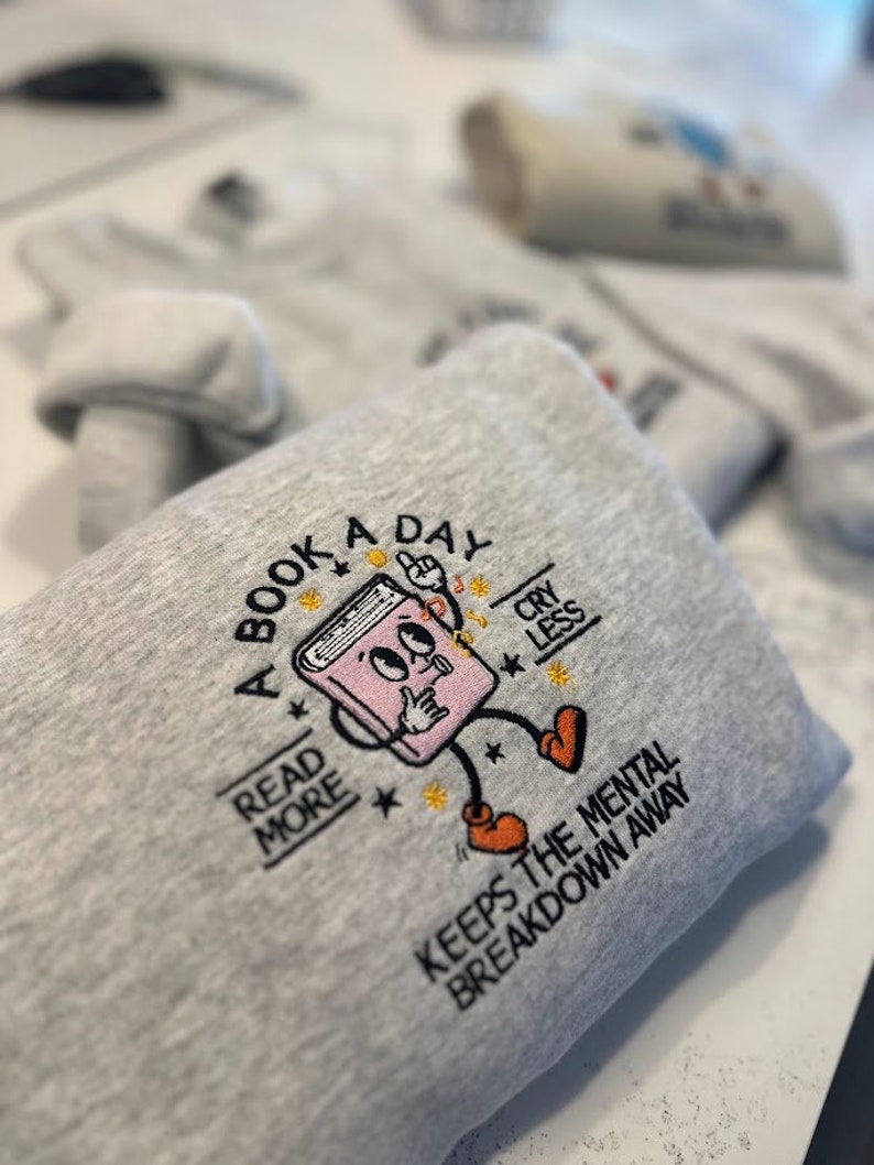 K&ouml;nnte beinhalten: Ein graues Sweatshirt mit einem gestickten Design eines Buches mit Beinen und Armen. Das Buch h&auml;lt sich einen Finger auf die Lippen und hat eine Gedankenblase &uuml;ber seinem Kopf. Der Text auf dem Sweatshirt lautet "A Book A Day Read More Cry Less Keeps The Mental Breakdown Away".