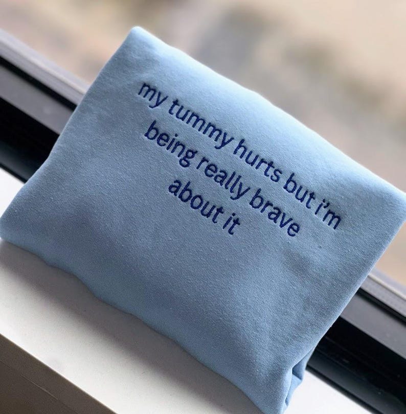 May include: Light blue sweatshirt with the text "my tummy hurts but i'm being really brave about it" embroidered in blue.