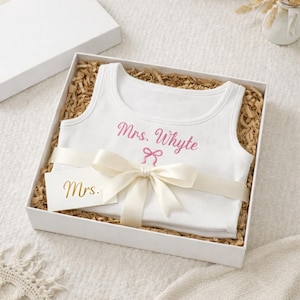 May include: A white tank top with the text "Mrs. Whyte" in pink script, presented in a white gift box with a cream ribbon and a small card that says "Mrs.". The box is filled with brown packing material.