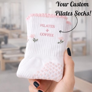Könnte beinhalten: Weiße Pilates-Socken mit rosa Akzenten und rutschfesten Griffen. Die Socken zeigen den Text "PILATES + COFFEE" und florale Stickereien. Der obere Rand ist mit einem rosa, gewellten Design versehen. Der Text "Your Custom Pilates Socks!" ist ebenfalls enthalten.