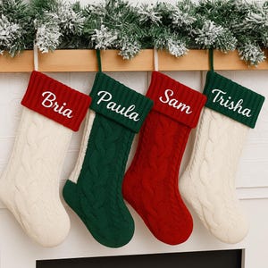 May include: Four personalised Christmas stockings in red, green, and cream colours hang from a wooden mantel. Each stocking has a cable knit design and a name embroidered on the cuff: Bria, Paula, Sam, and Trisha. Festive holiday decor.