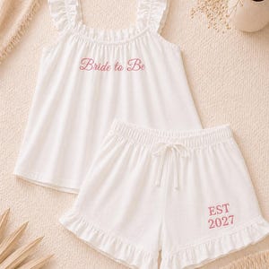 May include: White pajama set with a tank top and shorts. The tank top has ruffle straps and the words "Bride to Be" in pink. The shorts have a drawstring waist and the text "EST 2027" in pink. The set is made of a soft fabric.
