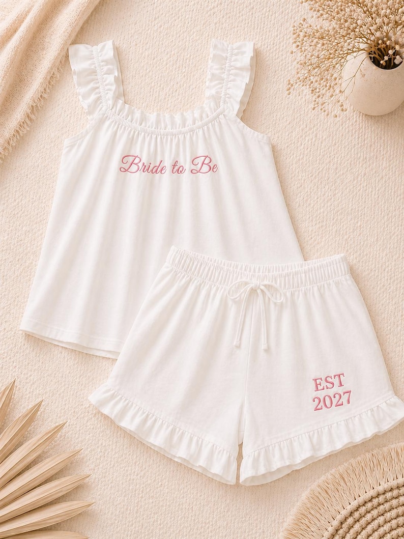 May include: White pajama set with a tank top and shorts. The tank top has ruffle straps and the words "Bride to Be" in pink. The shorts have a drawstring waist and the text "EST 2027" in pink. The set is made of a soft fabric.