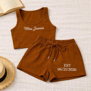 May include: A brown ribbed knit two-piece set. The crop top has "Mrs. James" in white script. The shorts have a drawstring waist and "EST 06/21/2026" in white. A straw hat and book are in the background.