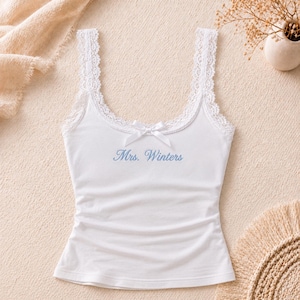 Puede incluir: Una camiseta de tirantes blanca con escote redondo, ribete de encaje blanco y un pequeño lazo de satén blanco en el centro. El texto Mrs. Winters está bordado en letra cursiva azul claro en la zona del pecho de la prenda de tirantes anchos.