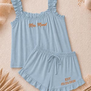 May include: A light blue pajama set featuring a tank top and shorts. The tank top has ruffled straps and the embroidered text "Mrs. Monet". The shorts have a drawstring waist and the embroidered text "EST. 05/25/2026".