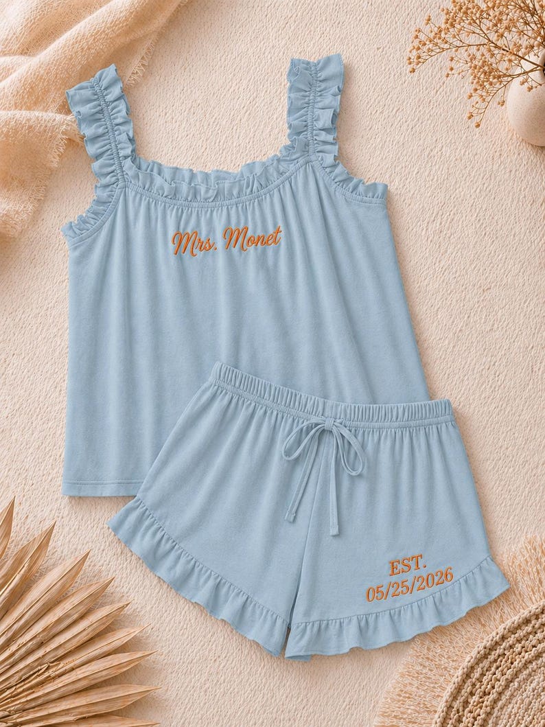 May include: A light blue pajama set featuring a tank top and shorts. The tank top has ruffled straps and the embroidered text "Mrs. Monet". The shorts have a drawstring waist and the embroidered text "EST. 05/25/2026".