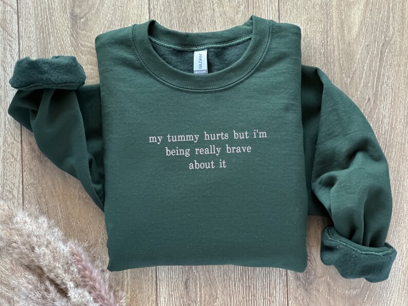 May include: A dark green sweatshirt with the text "my tummy hurts but I'm being really brave about it" in white.