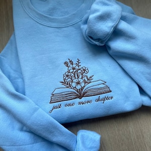 Peut inclure: Un sweat-shirt bleu clair avec un motif brodé marron d'un livre ouvert avec des fleurs sur le dessus. Le texte "just one more chapter" est brodé sous le livre.