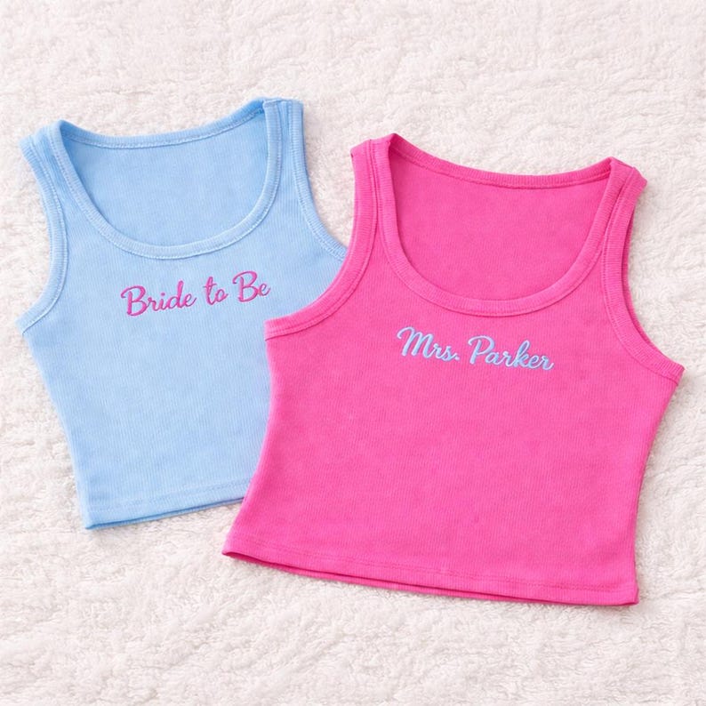 May include: Two tank tops, one light blue and one pink, are displayed. The light blue tank top has the embroidered text "Bride to Be" in pink. The pink tank top has the embroidered text "Mrs. Parker" in light blue.