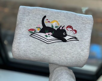 Sudadera con gato bordado sobre un libro, estampado floral de setas, ideal para amantes de los libros.