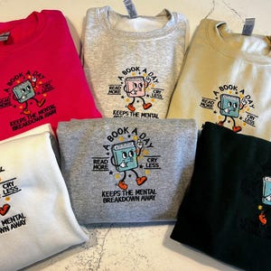 K&ouml;nnte beinhalten: F&uuml;nf Sweatshirts in verschiedenen Farben mit einer Grafik eines Buches mit einem Gesicht und dem Text "A Book A Day" mit den Worten "Read More" und "Cry Less" auf beiden Seiten des Buches. Der Text "Keeps the Mental Breakdown Away" befindet sich unter dem Buch.