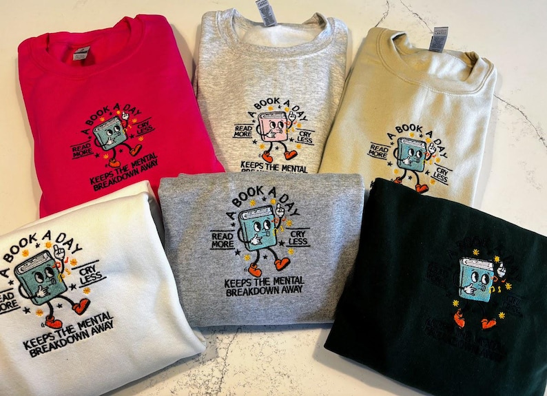 K&ouml;nnte beinhalten: F&uuml;nf Sweatshirts in verschiedenen Farben mit einer Grafik eines Buches mit einem Gesicht und dem Text "A Book A Day" mit den Worten "Read More" und "Cry Less" auf beiden Seiten des Buches. Der Text "Keeps the Mental Breakdown Away" befindet sich unter dem Buch.