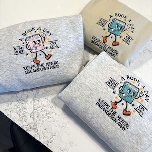 K&ouml;nnte beinhalten: Drei graue Sweatshirts mit einem blauen und orangefarbenen Stickmotiv eines Buches mit dem Text "A Book A Day" und "Read More, Cry Less, Keeps the Mental Breakdown Away".