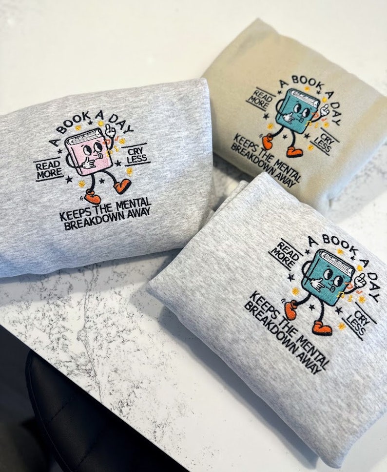 K&ouml;nnte beinhalten: Drei graue Sweatshirts mit einem blauen und orangefarbenen Stickmotiv eines Buches mit dem Text "A Book A Day" und "Read More, Cry Less, Keeps the Mental Breakdown Away".
