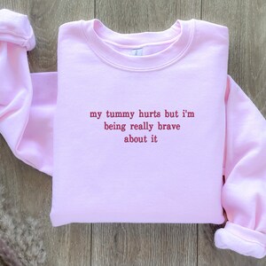 May include: Pink sweatshirt with the text "my tummy hurts but i'm being really brave about it" in red.