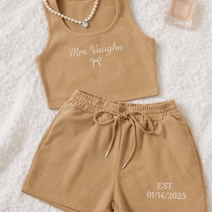 May include: Tan ribbed knit crop top and shorts set. The top is embroidered with "Mrs. Vaughn" and a bow. The shorts have a drawstring waist and the date "EST 01/14/2025". A pearl necklace and perfume bottle are also in the image.
