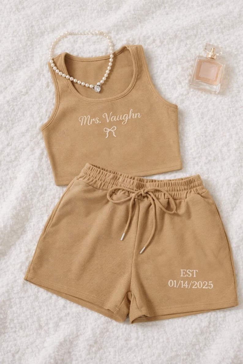 May include: Tan ribbed knit crop top and shorts set. The top is embroidered with "Mrs. Vaughn" and a bow. The shorts have a drawstring waist and the date "EST 01/14/2025". A pearl necklace and perfume bottle are also in the image.