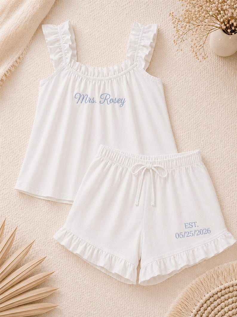 May include: White pajama set with a ruffled top and shorts. The top is embroidered with "Mrs. Rosey" in blue script. The shorts have a drawstring waist and are embroidered with "EST. 05/25/2026". The set is made of a soft fabric.